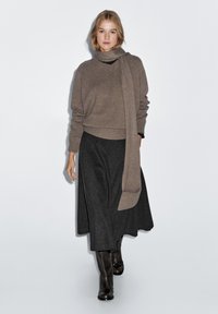 Brauner Strickpullover mit hohem Kragen, kombiniert mit einem schwarzen gemusterten Rock. Das Outfit wird durch kniehohe braune Stiefel vervollständigt.