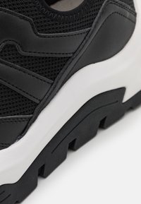 Černá sneaker s texturami z materiálů mesh a kůže, vybavená robustní bílou mezipodešví a černou podešví s drážkováním, zdůrazňující moderní designové prvky.