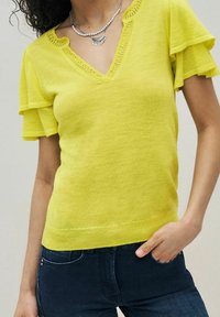 Gul strikket top med V-hals, blondekant og voluminøse kortærmer. Båret med mørkeblå jeans, hvilket giver et afslappet udseende.