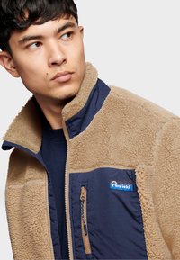 Fleece jacket with beige and navy panels, featuring a high collar, zippered pockets, and brand logo on the chest. Soft texture and warm design.