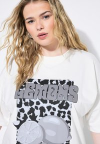 Camiseta blanca de gran tamaño con un diseño gráfico que presenta la palabra "LEMONS" en gris, sobre un fondo de estampado animal en blanco y negro.