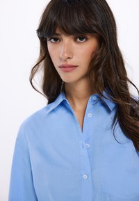 Camicia azzurra chiaro con bottoni, realizzata in tessuto liscio, dotata di colletto appuntito e taschino sul lato sinistro.