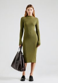 Olivengrünes, geripptes Langarmkleid mit hohem Ausschnitt, kombiniert mit einer dunkelbraunen, glänzenden Umhängetasche und schwarzen Slippern.