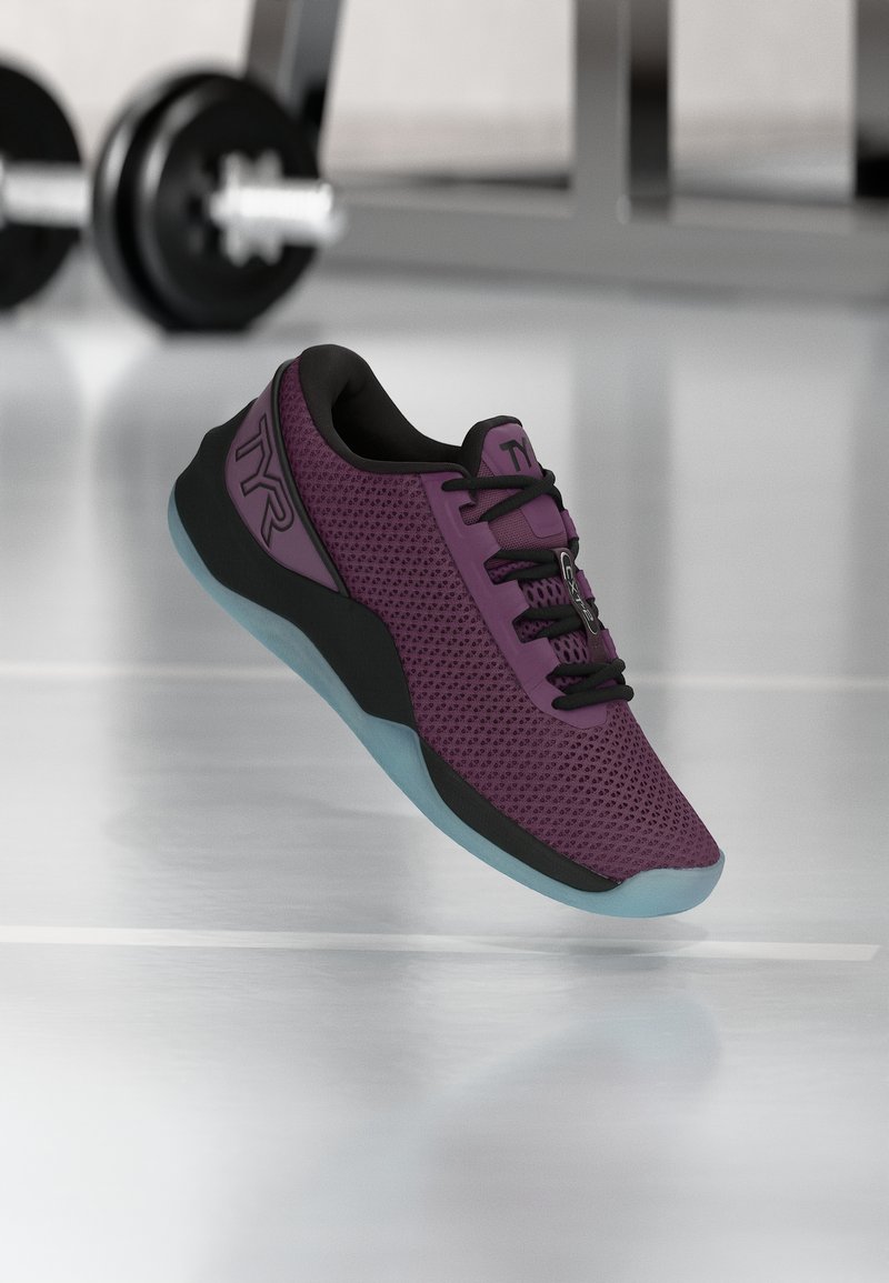 Αθλητικό παπούτσι σε μοβ και μαύρο με λογότυπο TYR και ανοιχτό μπλε σόλα, τοποθετημένο στον αέρα πάνω από το παρκέ γυμναστηρίου με αλτήρα στο φόντο.