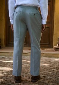 Hellegraue, maßgeschneiderte Hose mit flacher Front, zwei Gesäßtaschen und dezenten Gürtelschlaufen. Glatte Stoffoberfläche, kombiniert mit braunen Stiefeletten.