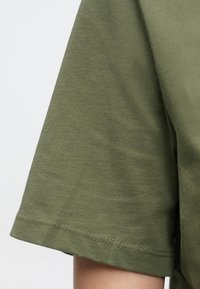 Olīvu zaļš kokvilnas T-krekls ar īsām piedurknēm, kas redzams tuvplānā uz personas rokas pret vienkārša gaiša fona.