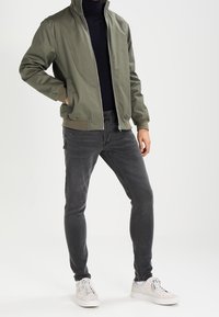 Olivgrön bomberjacka med hög krage, dragkedja, kombinerad med mörkgrå slim-fit jeans och vita sneakers. Mjukt tyg.