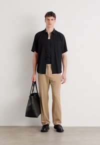 Camicia nera a maniche corte con bottoni, t-shirt nera, pantaloni beige, scarpe nere, con una borsa tote nera. Outfit minimalista contro uno sfondo semplice.
