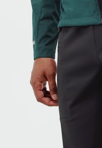 Right hand relaxed beside torso, wearing dark pants and green long-sleeve jacket, against a plain light background.