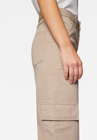 Beige Cargo-Hosen aus strapazierfähigem Stoff, mit einer Seitentasche und sichtbaren Nähten. Der Bund ist mit einem Gürtelschlaufe ausgestattet.