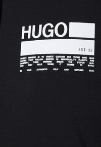 Tessuto nero con scritta bianca in grassetto "HUGO", "EST 93" e una dichiarazione di intenti sulla individualità e autenticità nel design dell'abbigliamento.