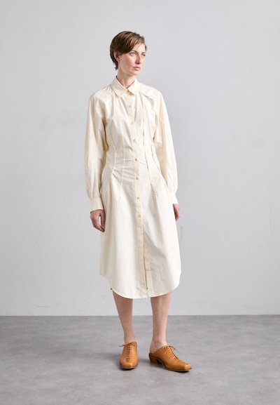 Robe chemise longue de couleur crème à manches longues avec des boutons sur le devant, des coutures ajustées et un ourlet arrondi, associée à des chaussures à lacets brunes.