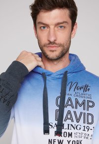 Felpa con cappuccio caratterizzata da una gradazione ombre che va dal blu al bianco, dettagli neri sulle maniche e stampa di testo sul davanti.