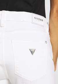 Białe dżinsy z srebrnym trójkątnym logo na tylnej kieszeni oraz markową metką w talii. Gładka tekstura i dopasowany krój.