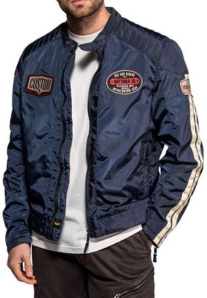 Un modelo masculino aparece parcialmente encuadrado con una chaqueta azul marino con cremallera, parches de carreras y rayas color crema en las mangas, sobre una camisa blanca y pantalones oscuros.