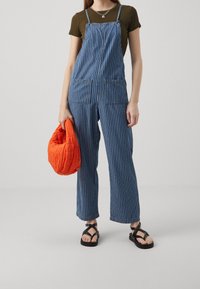 Nainen, jolla on päällään sinivalkoiset raidalliset farkkuhaalarit, oliivinvihreä t-paita, mustat sandaalit ja hän pitää kirkkaan oranssia tilkkulaukkua.