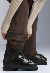 Černé lyžařské boty s nastavitelnými přezkami, doplněné hnědými lyžařskými kalhotami s elastickými manžetami a hladkým, mírně lesklým povrchem.