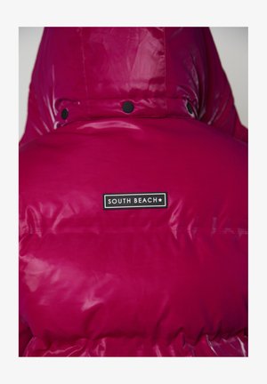 Chaqueta acolchada fucsia con una textura brillante y esponjosa y capucha. Presenta una etiqueta negra con el texto "SOUTH BEACH". Broches cerca del cuello.