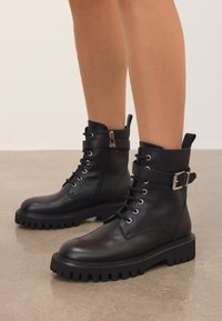 Botine negre din piele, cu talpă groasă, șireturi, fermoar lateral și curea cu cataramă, purtate de o persoană care stă pe un paviment lustruit.