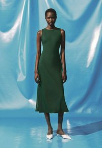Abito midi verde senza maniche con una silhouette aderente. Indossato con scarpe bianche e collant a trama su uno sfondo blu.