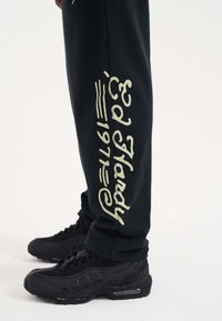 Svarta sweatpants i bomull med en lös passform, prydda med vit kursiv text på sidan. Matchas med svarta sneakers för kontrast.