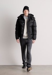 Μαύρο puffer μπουφάν με ψηλό γιακά, επενδεδυμένο με κρεμ πουλόβερ και ανοιχτό μπλε πουκάμισο. Γκρι παντελόνι και μαύρα παπούτσια ολοκληρώνουν την εμφάνιση.