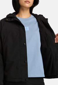 Svart huva jacka med tryckknappar, som bärs över en ljusblå t-shirt med en liten logotyp. Slät tyg med en avslappnad passform.