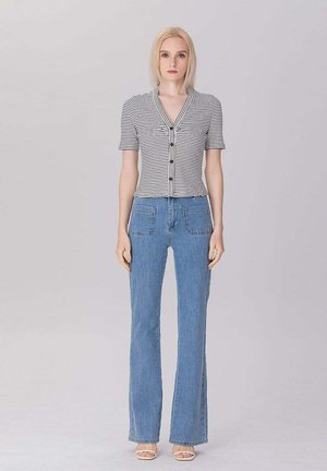 Jeune femme aux cheveux blonds et raides portant une chemise rayée à manches courtes boutonnée, un jean bleu avec des poches avant et des sandales blanches.