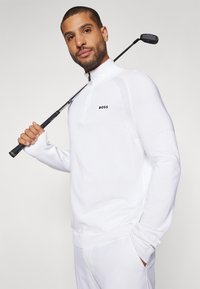 Bela zip-up golf pulover iz teksturirane tkanine, ki ima črn logotip na prsih in rebraste poudarke na ramenih in rokavih.