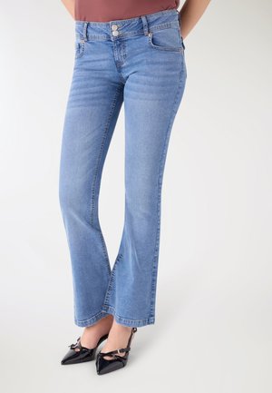 Donna che indossa jeans a vita alta azzurri con fondo svasato e décolleté slingback nere con punta a punta, in piedi contro uno sfondo bianco semplice.