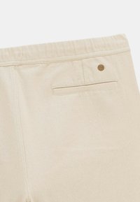 Vue arrière de pantalons beiges présentant une poche passepoilée unique avec un petit logo métallique rond au-dessus de la poche sur le côté droit.