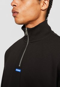 Black high-neck sweatshirt featuring a quarter zipper and a blue "HUGO" logo patch on the front. Smooth texture, casual design.
