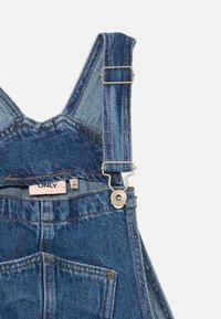 Pechera de mezclilla azul con tirantes ajustables, hebilla de metal y botón, bolsillo frontal y etiqueta de marca interior "ONLY" sobre un fondo blanco.