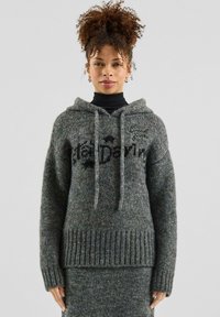 Woman with curly hair wearing a grey knit hooded sweater and matching skirt, standing against a plain light background.