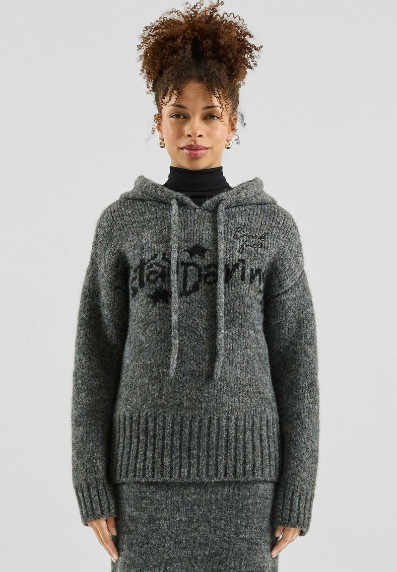 Woman with curly hair wearing a grey knit hooded sweater and matching skirt, standing against a plain light background.