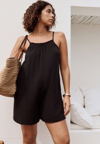 Tuta nera con trama, con spalline sottili e scollo arricciato, abbinata a una borsa a tracolla beige intrecciata.