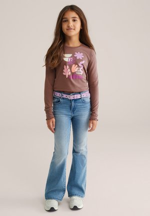 Chemise marron à manches longues avec un graphisme coloré et du texte, associée à un jean bleu évasé et une ceinture rose. Baskets aux tons clairs et foncés.