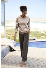 Light beige sweatshirt with "LONDON" stitched in a darker beige. Paired with dark ribbed trousers. Model walking barefoot on wooden deck.