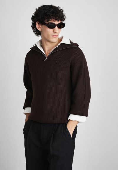 Brauner gerippter Pullover mit einem Reißverschlusskragen, kombiniert mit einem weißen Hemd mit Kragen. Model trägt schwarze Sonnenbrille und schwarze Hosen.
