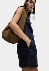 Haut marine avec des détails à lacets associé à une jupe marine. Le mannequin tient un sac marron au toucher doux et décontracté contre un fond blanc uni.