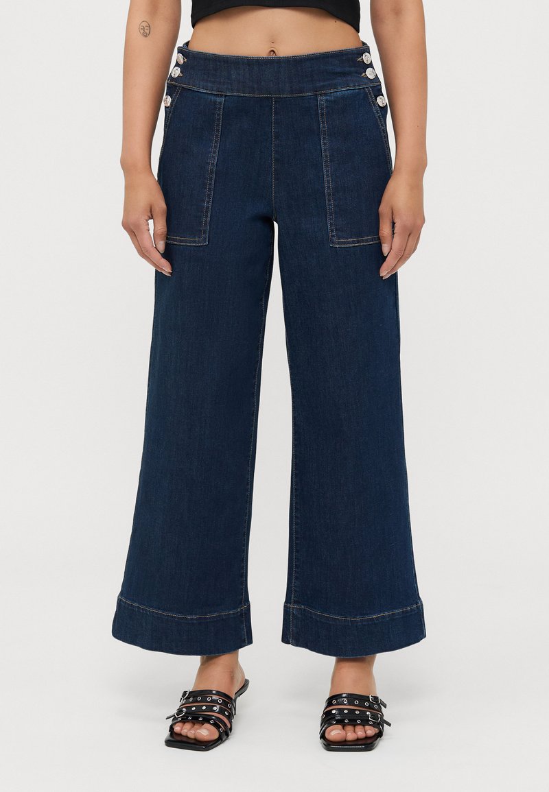 Pantalones vaqueros de pierna ancha en azul oscuro con una cintura alta, que presentan grandes bolsillos frontales y acentos decorativos de botones a los lados.