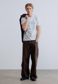 Ανοιχτό γκρι μπλουζάκι με την επιγραφή "BOSS" σε πιο σκούρο γκρι, συνδυασμένο με καφέ παντελόνια φαρδιάς γραμμής και μαύρα παπούτσια, κρατώντας ένα σκούρο σακάκι.