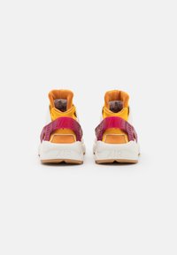 Ett par vita och orange Nike Air-sneakers med vinröda hälremmar, sedda bakifrån mot en enfärgad ljus bakgrund.