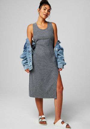 Woman wearing a sleeveless gray midi dress with a side slit, holding a blue denim jacket off her shoulders, and white buckle sandals.