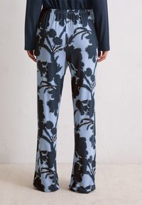 Personne portant un pantalon ample avec de grands motifs floraux noirs sur fond bleu clair, se tenant contre un mur uni.