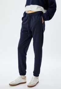 Marineblauwe sweatpants met elastische boorden en een drawstring taille, voorzien van zijzakken en een gladde textuur. Gecombineerd met lichte sneakers.