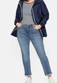 Giacca a vento navy con rifiniture bianche, t-shirt a righe, jeans azzurri con risvolti, e sneakers bianche. La giacca ha una chiusura a zip e tasche.