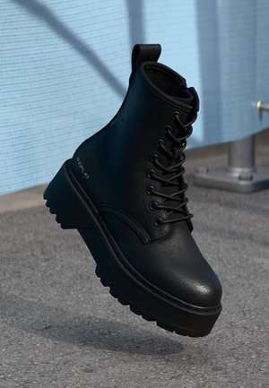 Schwarzer Leder-Schnürstiefel mit Plateausohle und dickem Profil, mit der Marke „REPLAY“, schwebt über einer Asphaltfläche nahe einer hellblauen Wand und einem Metallpfosten.