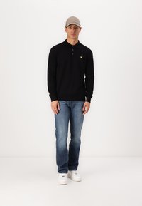 Polo shirt negru cu mânecă lungă, cu închidere cu nasturi, asortat cu blugi albaștri și adidași albi. O șapcă bej completează ținuta. Logo galben pe cămașă.