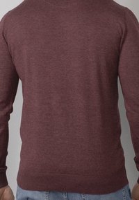 Uomo che indossa un maglione lungo a maniche semplici di color bordeaux e jeans azzurri chiari, visto da dietro su uno sfondo grigio.